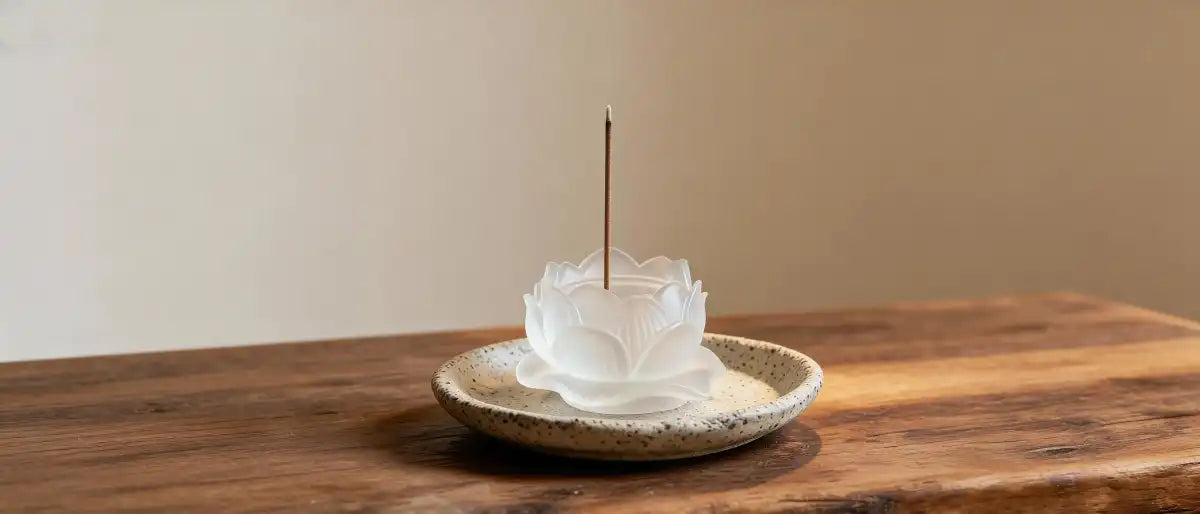 A white, frosted glass lotus flower-shaped incense burner sitting on a speckled beige ceramic plate on a wooden table, with a single incense stick burning