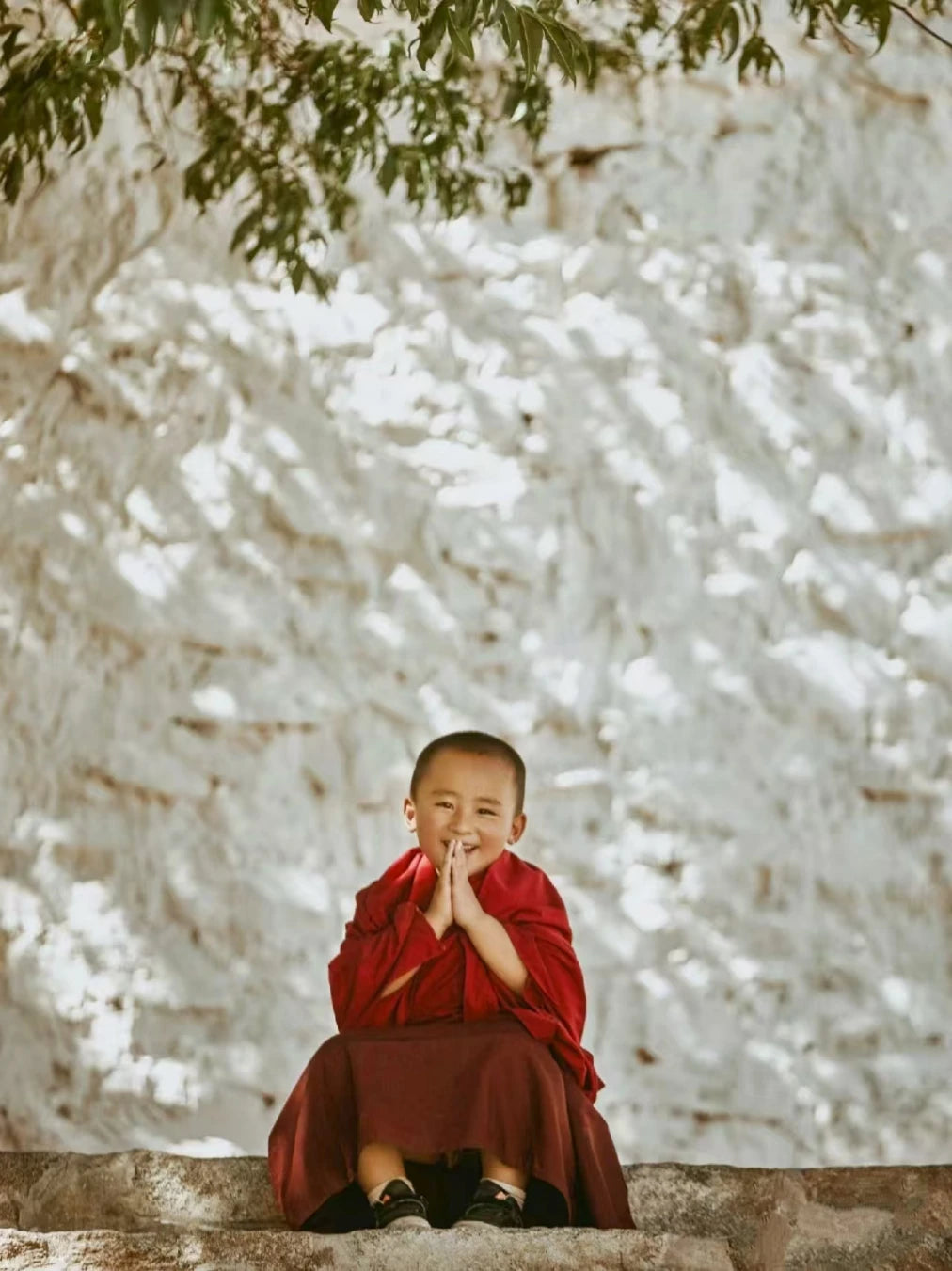 Child in red robe sitting on a stone ledge with a textured white wall and greenery in the background