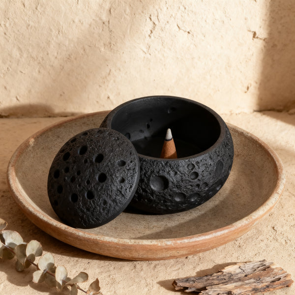 Black ceramic incense holder with a small candle on a beige plate, set against a neutral background.