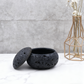 Black textured pot with lid on a marble surface with a geometric vase in the background.