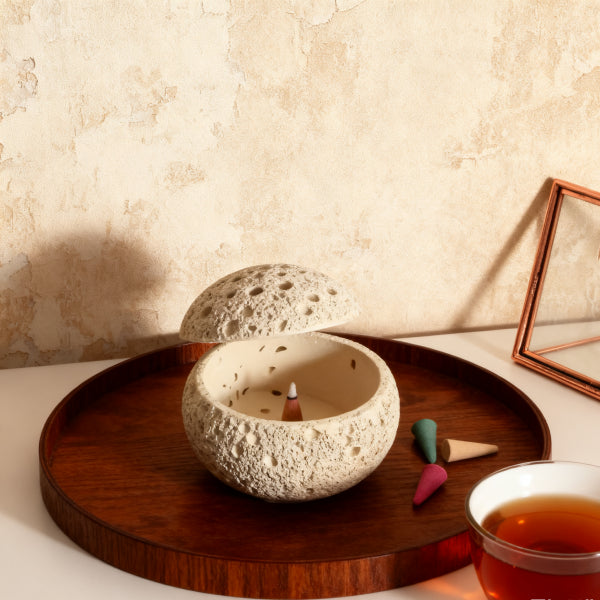 Ceramic incense burner on a wooden tray with tea and decorative items against a beige wall.