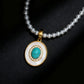 Close-up product shot of the Balance Grace necklace against a black background, featuring a white pearl strand and a gold oval pendant with a central turquoise stone surrounded by a crystal halo.