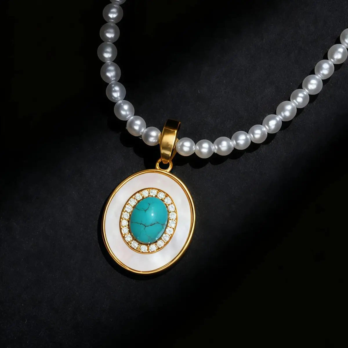 Close-up product shot of the Balance Grace necklace against a black background, featuring a white pearl strand and a gold oval pendant with a central turquoise stone surrounded by a crystal halo.