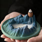 Hands holding Tibetan Backflow Incense Burner with Mountain Design, Cone Incense, and Flowing Smoke on Dark Background