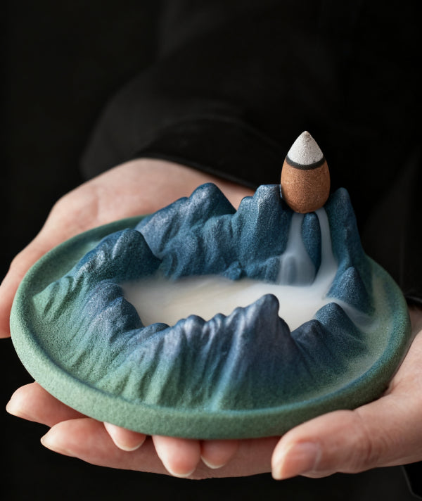 Hands holding Tibetan Backflow Incense Burner with Mountain Design, Cone Incense, and Flowing Smoke on Dark Background
