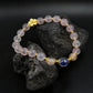 The Healing & Abundance bracelet resting on a dark, porous rock. The contrast highlights the translucency of the pink "Blue Core Agate" beads and the shimmering gold inclusions within the blue "Ash-Liuli" bead, symbolizing grounded energy.
