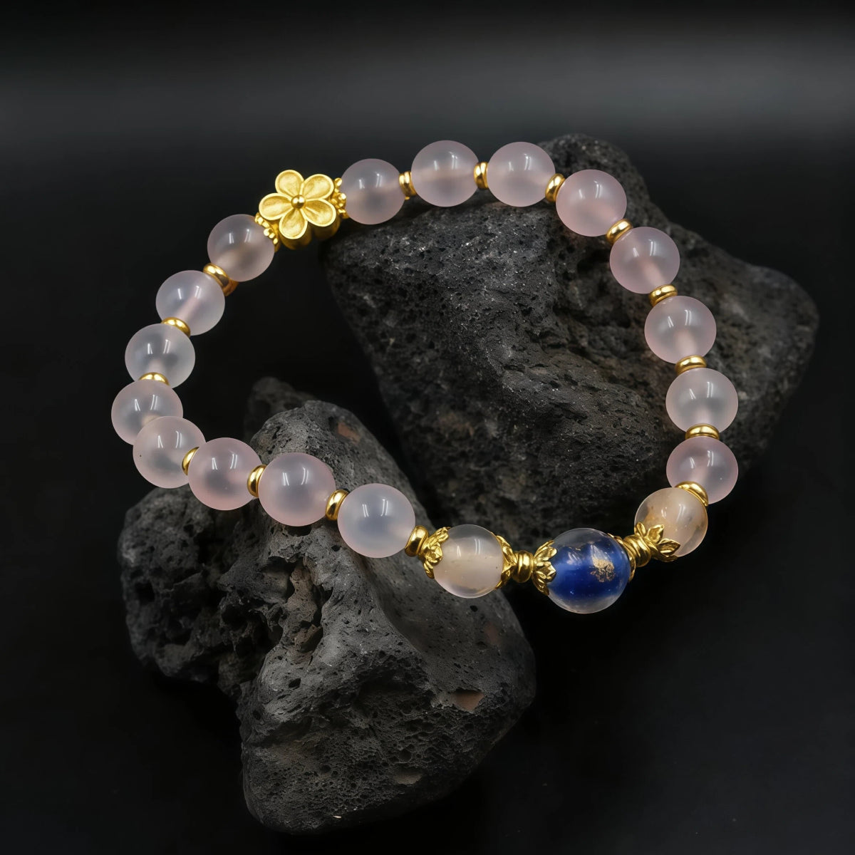 The Healing & Abundance bracelet resting on a dark, porous rock. The contrast highlights the translucency of the pink "Blue Core Agate" beads and the shimmering gold inclusions within the blue "Ash-Liuli" bead, symbolizing grounded energy.