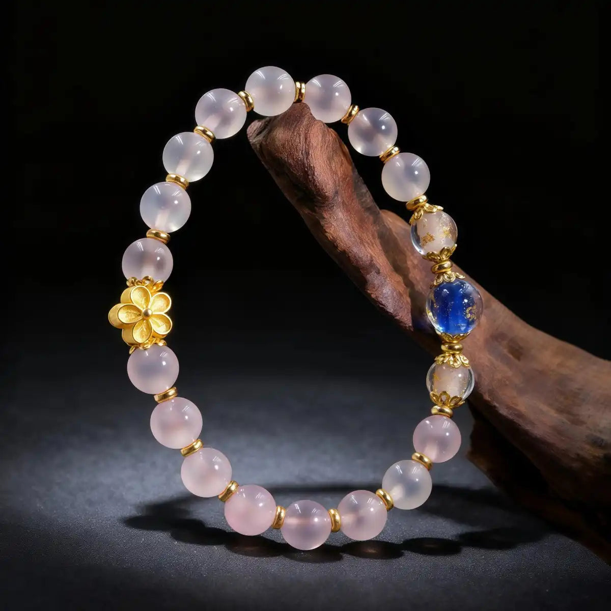 The "Healing & Abundance" bracelet draped vertically over a rustic wooden branch. It features translucent pink "Blue Core Agate" beads, a central "Blue & Gold Foil Ash-Liuli" bead for wealth, and an 18k gold-plated flower charm.