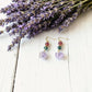 Pair of earrings with purple stones on a white wooden surface with lavender flowers.