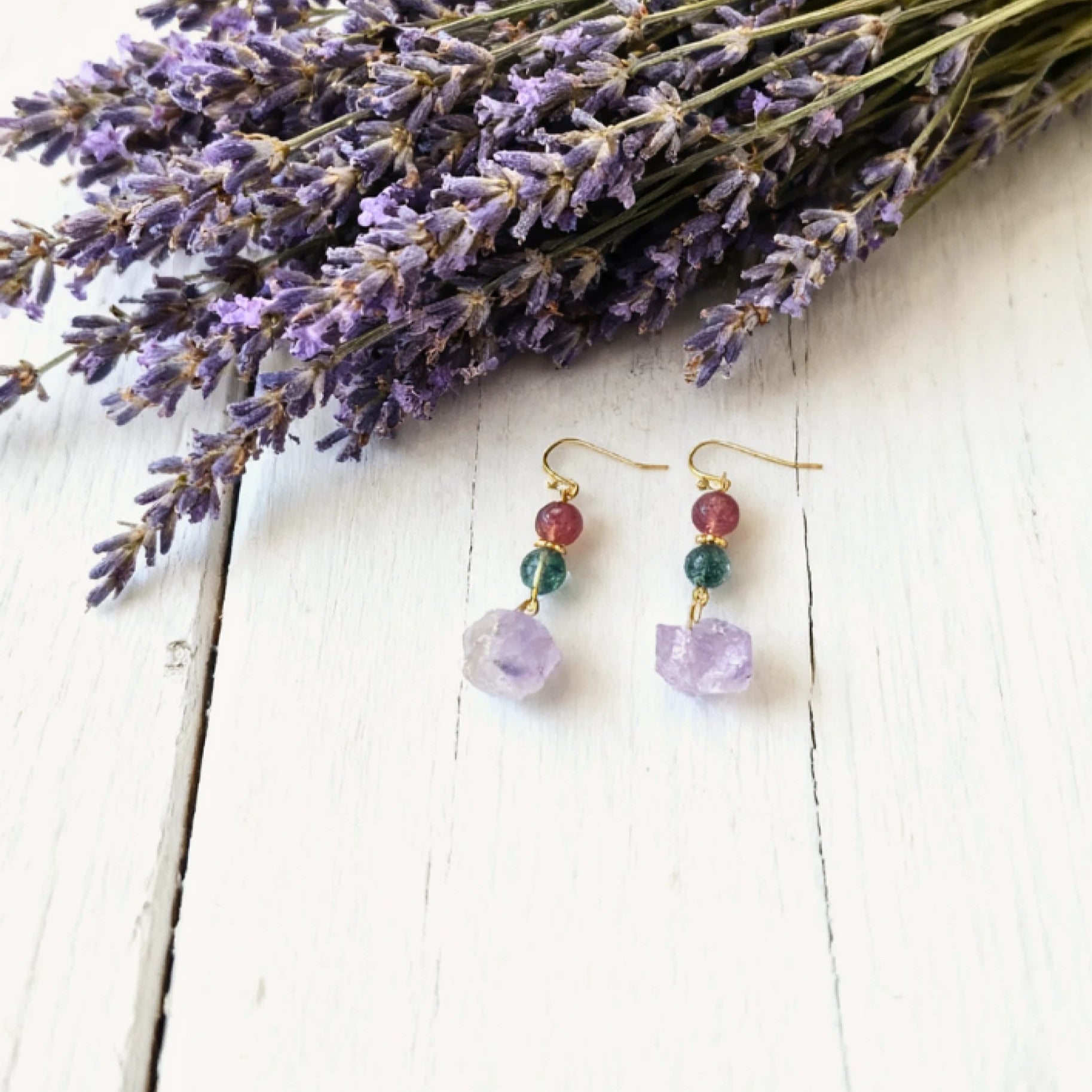 Pair of earrings with purple stones on a white wooden surface with lavender flowers.