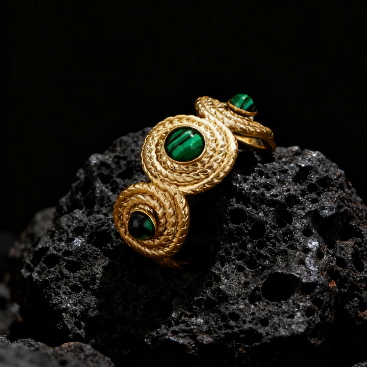A close-up side view of the gold Five Elements Malachite ring resting on volcanic rock, showcasing the intricate spiral pattern on the metalwork and the detail of the central green Malachite setting.
