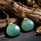 A pair of Tibetan Amazonite earrings resting on a piece of textured driftwood. The natural light highlights the geometric faceting and white inclusions of the green stone, evoking a sense of nature and renewal.