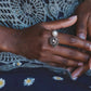 Close-up of a hand wearing a decorative ring with a pearl, against a textured fabric background.