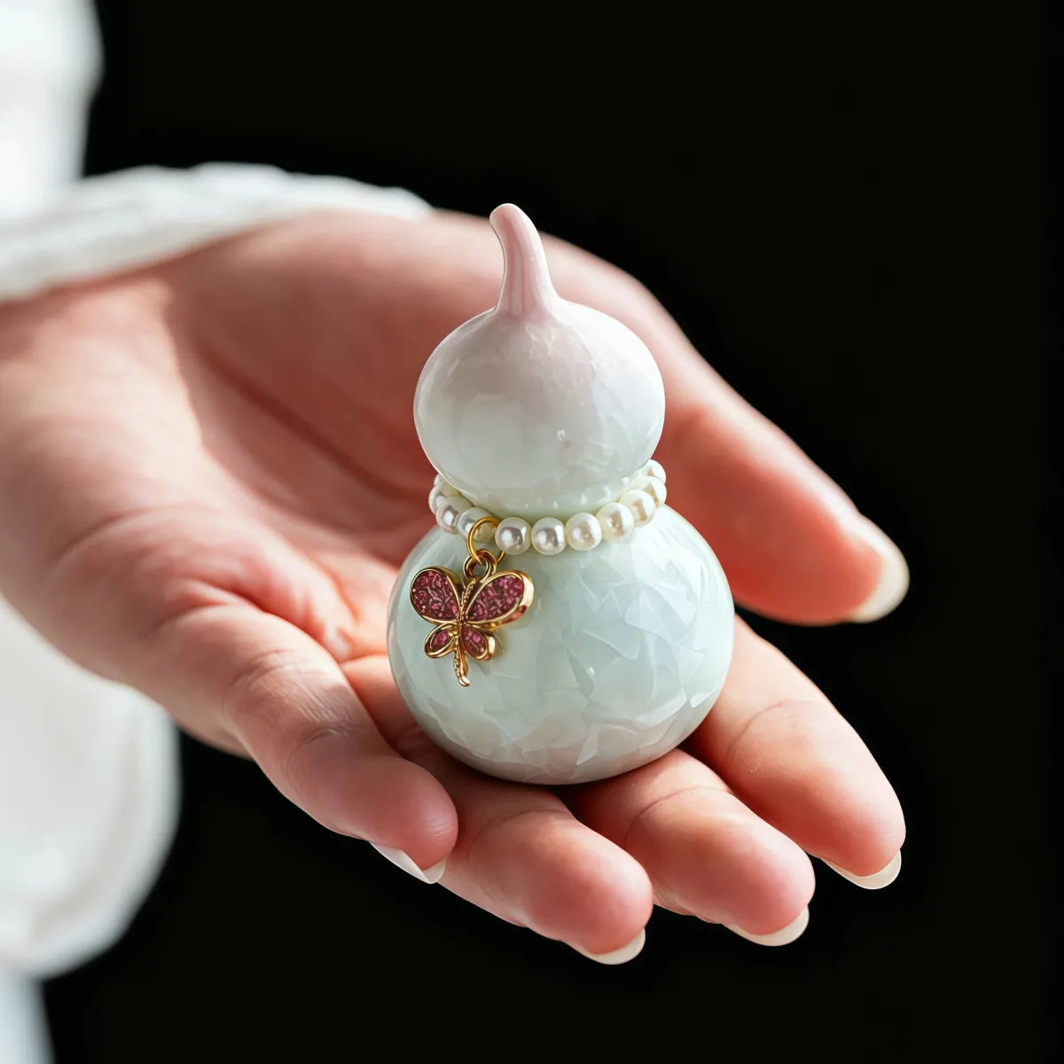 Hand holding a Feng Shui Pearl Gourd Ornament with a pearl bracelet and butterfly charm against a black background