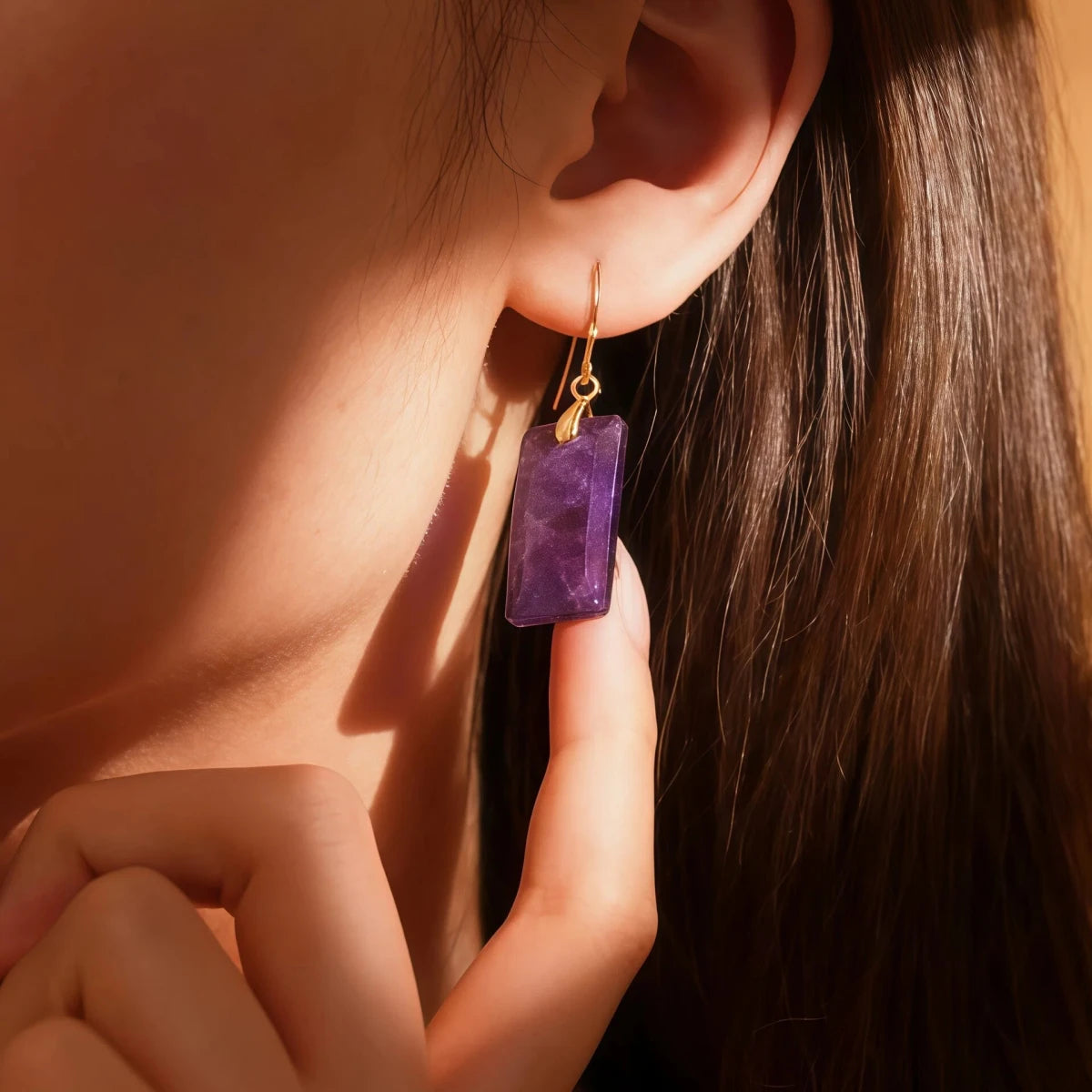 A close-up profile of a woman wearing rectangular Tibetan Amethyst earrings. The deep purple gemstone contrasts elegantly with her skin, representing a talisman for wisdom, intuition, and the Crown Chakra.