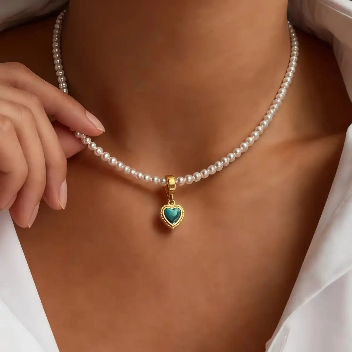A woman in a white shirt wearing a delicate seed pearl necklace featuring a gold-rimmed, heart-shaped Tibetan turquoise pendant. Her hand gently touches the necklace, demonstrating its elegant fit and style.
