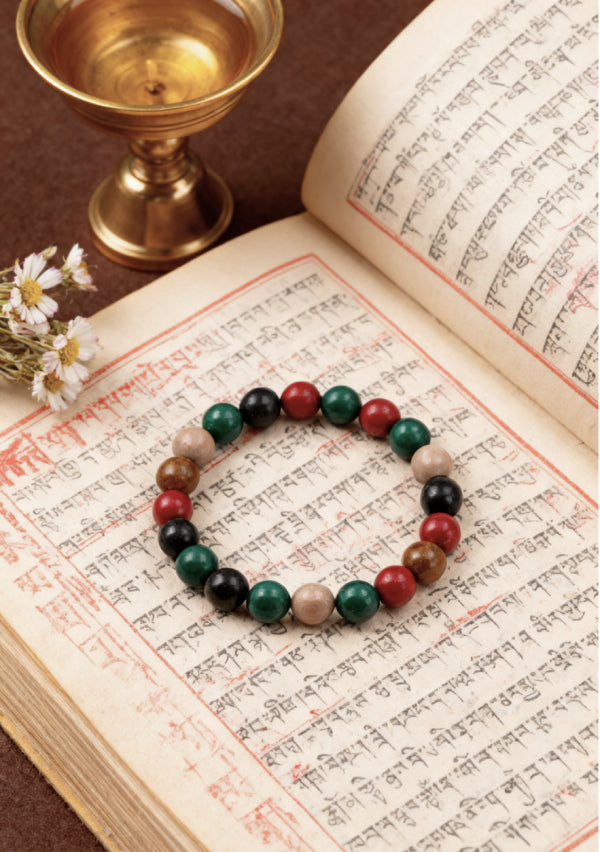 Handmade stretch bracelet with black, red, green, and natural wooden beads displayed on ancient manuscript with brass oil lamp and daisies.