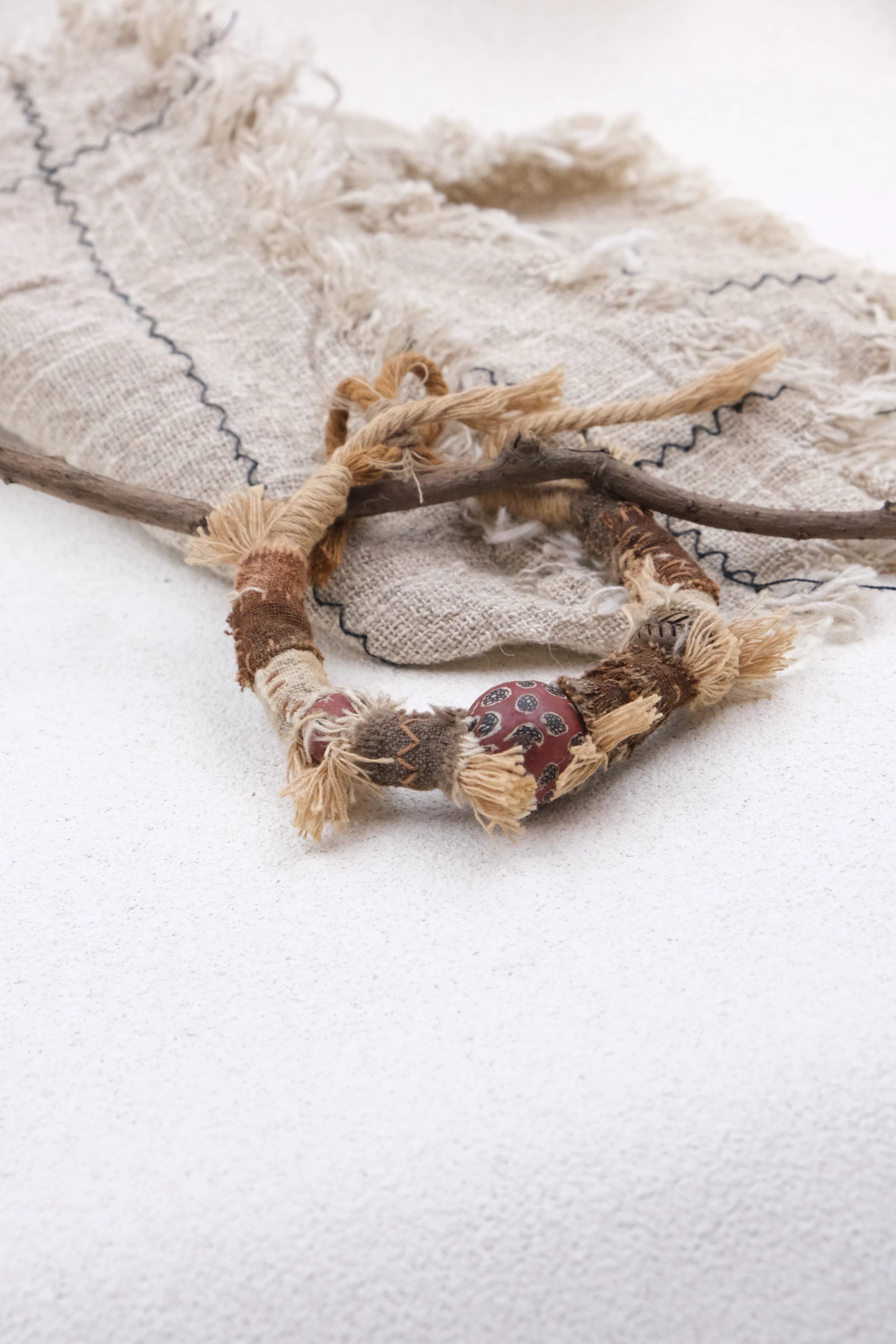 Handcrafted Ancient Guardian Bracelet made of braided natural fibers with frayed tassel details, featuring a central red ancient glass charm, decorated with wood and fabric