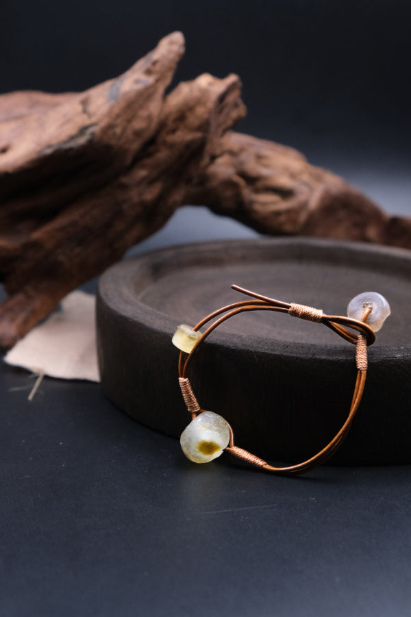 Unique Sun Spirit Bracelet adorned with ancient Liuli beads, strung on brown leather cord, resting on a dark - toned wooden disc, with textured, weathered wood in the background, promoting grounding and inner clarity. 