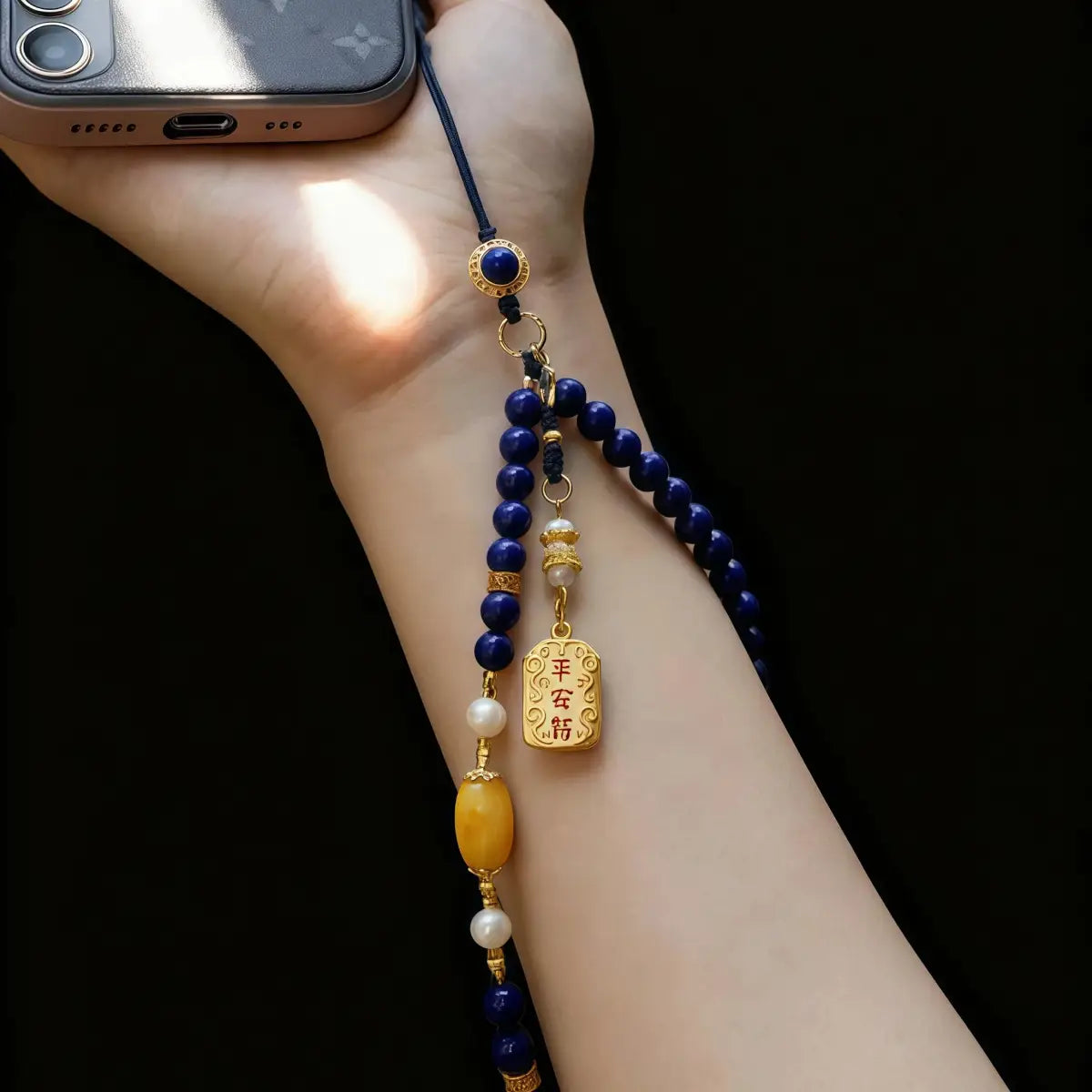 Hand holding a beaded phone charm with blue, gold, and pearl beads against a black background