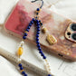 Phone charm with blue beads and a gold tag on a phone case.