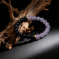 The Tibetan Dual Energy Bracelet draped over a piece of rustic driftwood. The lighting highlights the clarity of the amethyst and the sheen of the black obsidian, evoking a sense of natural healing and spiritual connection.