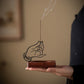 Hand holding a wooden incense holder with a hand design and smoking incense stick.