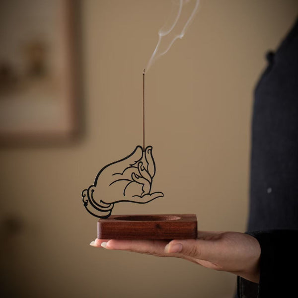 Hand holding a wooden incense holder with a hand design and smoking incense stick.