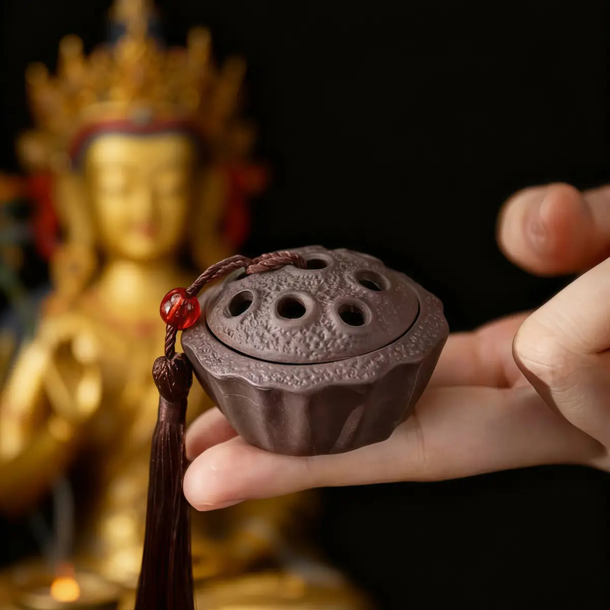 Hand holding a brown tibetan incense burner with a blurred golden statue in the background