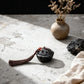 Black decorative item with tassel on a textured surface with a vase and rocks in the background