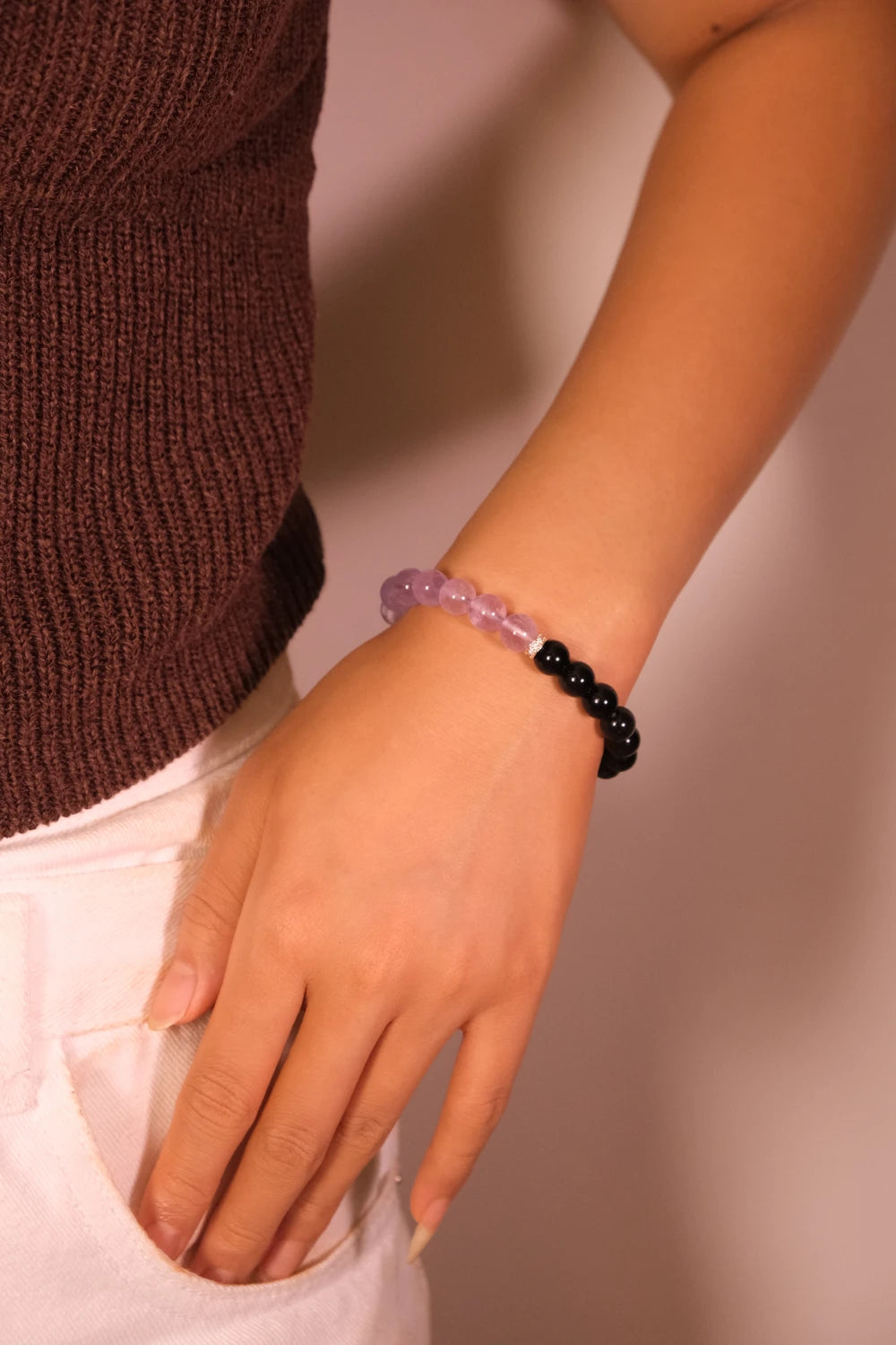 Close-up of a woman wearing the Tibetan Dual Energy Bracelet made of black obsidian and purple amethyst beads, photographed against a light pink background.