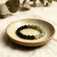 Bracelet with black and green beads on a ceramic dish with eucalyptus leaves in the background.