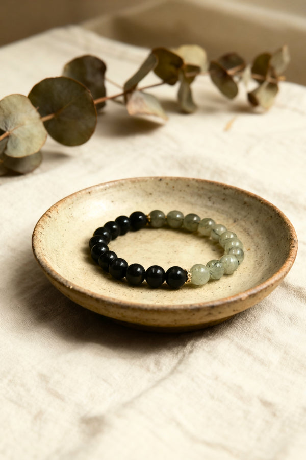 Bracelet with black and green beads on a ceramic dish with eucalyptus leaves in the background.