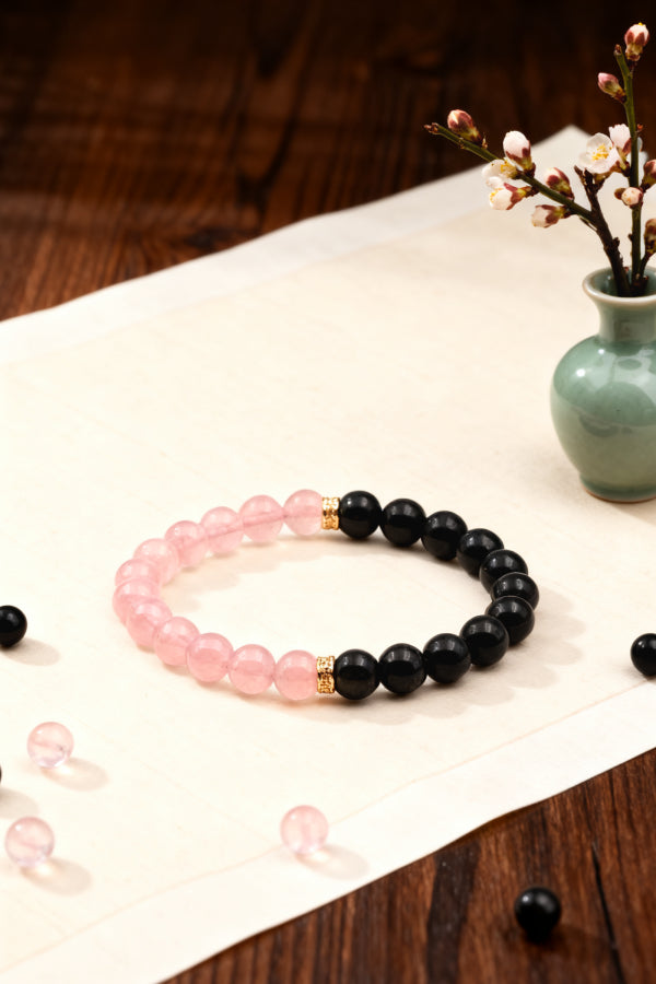 Two beaded bracelets, one pink and one black, on a wooden surface with a vase of flowers in the background.