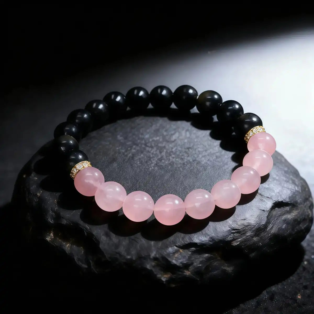 The "Love & Harmony" Tibetan Dual Energy Bracelet resting on a dark textured rock. It features a split design of black obsidian and pink rose quartz beads separated by gold accents, symbolizing the balance of protection and attracting love.