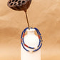 White ceramic vase wrapped with handmade multi-strand beaded necklace featuring blue beads, red spacers, and white round beads, styled with dried lotus pod decor.