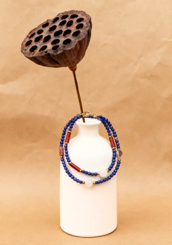 White ceramic vase wrapped with handmade multi-strand beaded necklace featuring blue beads, red spacers, and white round beads, styled with dried lotus pod decor.