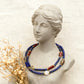 Artisan beaded necklace featuring lapis lazuli, red stone beads, gold spacers, and white pendant on classical marble bust jewelry display.