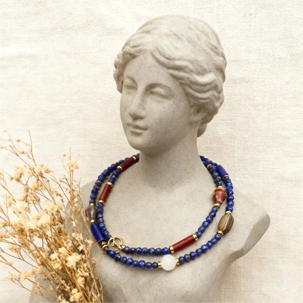 Artisan beaded necklace featuring lapis lazuli, red stone beads, gold spacers, and white pendant on classical marble bust jewelry display.