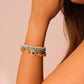 Close-up of a woman's arm wearing a bracelet featuring multiple strands with elements like pearls, turquoise, and ornate decorative beads on a beige background