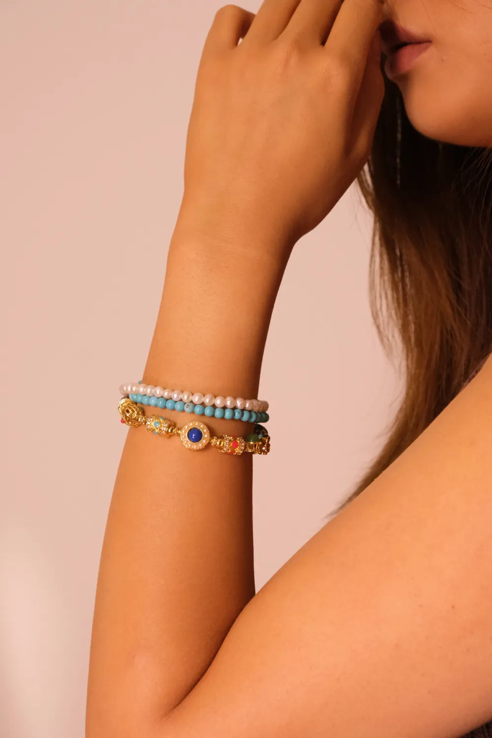 Close-up of a woman's arm wearing a bracelet featuring multiple strands with elements like pearls, turquoise, and ornate decorative beads on a beige background