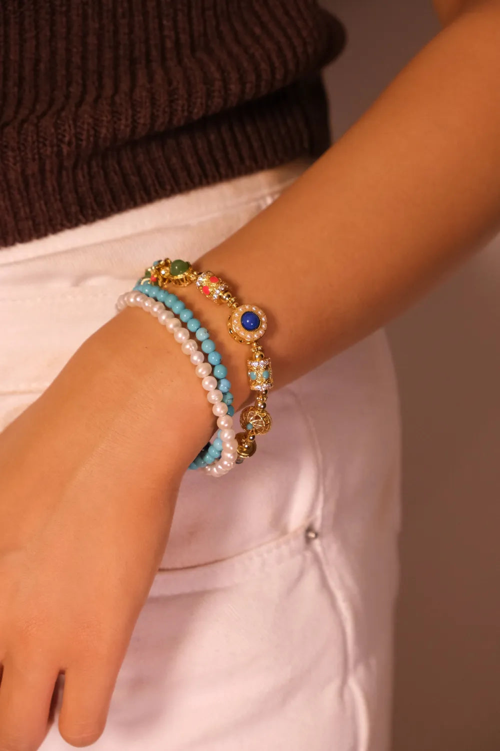 Close-up of a wrist wearing a bracelet featuring multiple strands with elements like pearls, turquoise, and ornate decorative beads on a neutral background
