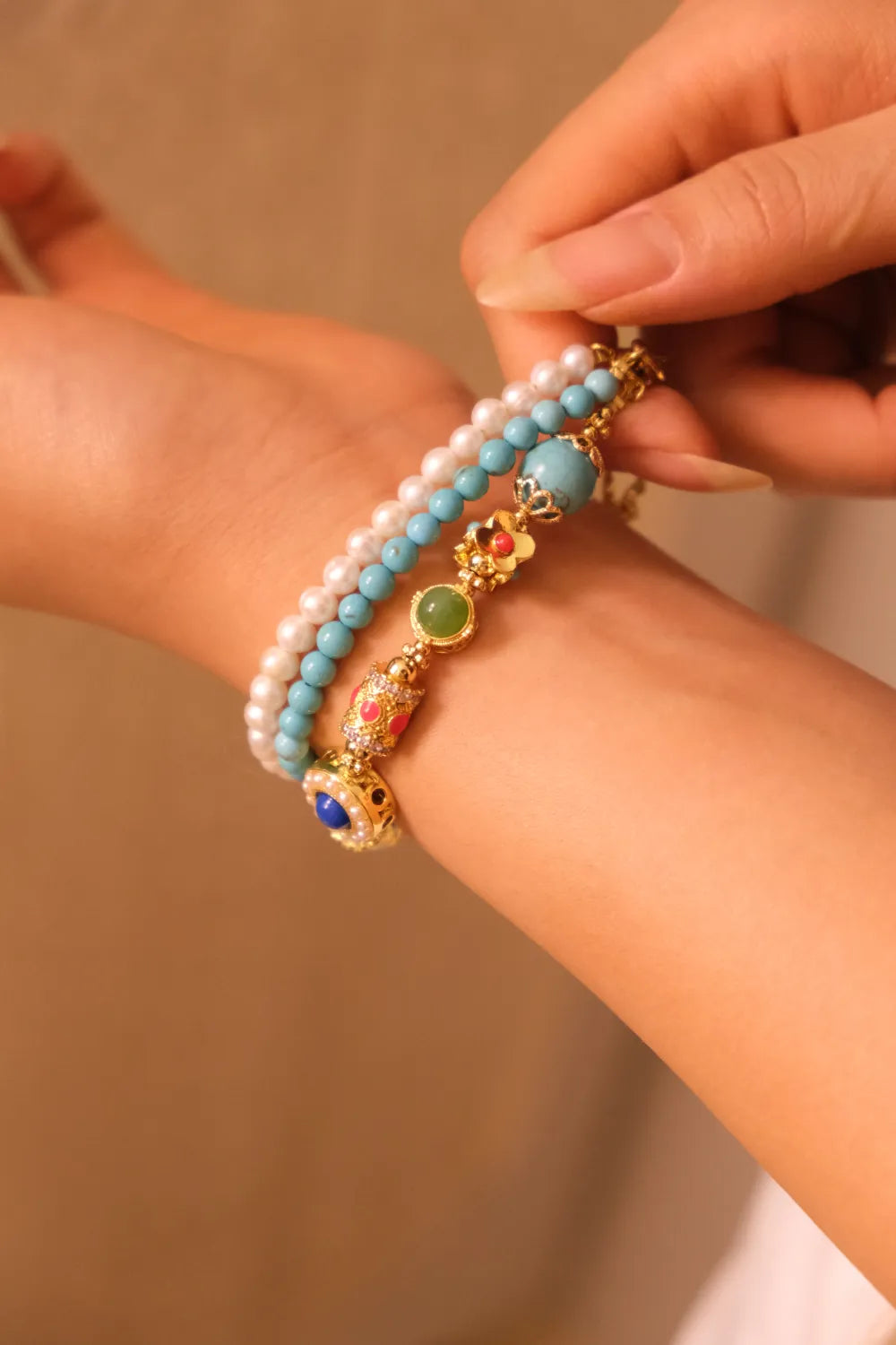 Close-up of a woman's arm wearing a bracelet featuring multiple strands with elements like pearls, turquoise, and ornate decorative beads on a beige background