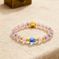 A bracelet featuring frosted pink beads, adorned with a golden clover charm, a vibrant blue bead, and translucent accent beads, placed on a beige textured fabric with an antique brass box in the background.
