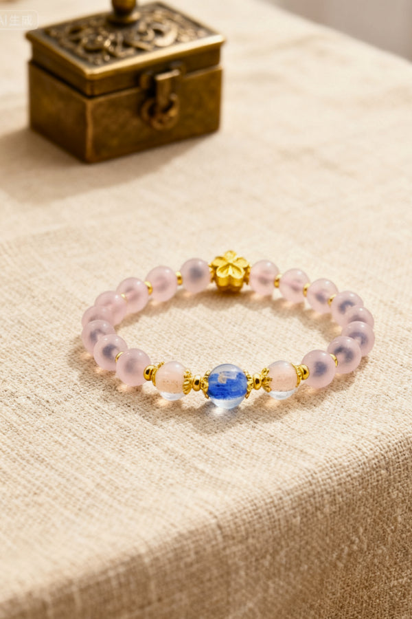 A bracelet featuring frosted pink beads, adorned with a golden clover charm, a vibrant blue bead, and translucent accent beads, placed on a beige textured fabric with an antique brass box in the background.