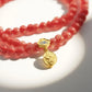 A double - stranded Tibetan Five Elements Flow Bracelet crafted with vibrant red foil ash - glass beads. It features a golden temple charm, adorned with a small decorative piece that has blue inlays. Hanging from the charm are two red beads. This bracelet is thought to promote vitality, attract abundance, and offer spiritual protection. 