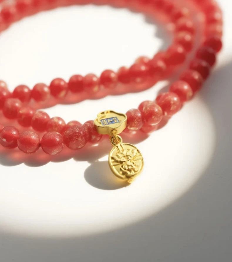 A double - stranded Tibetan Five Elements Flow Bracelet crafted with vibrant red foil ash - glass beads. It features a golden temple charm, adorned with a small decorative piece that has blue inlays. Hanging from the charm are two red beads. This bracelet is thought to promote vitality, attract abundance, and offer spiritual protection. 