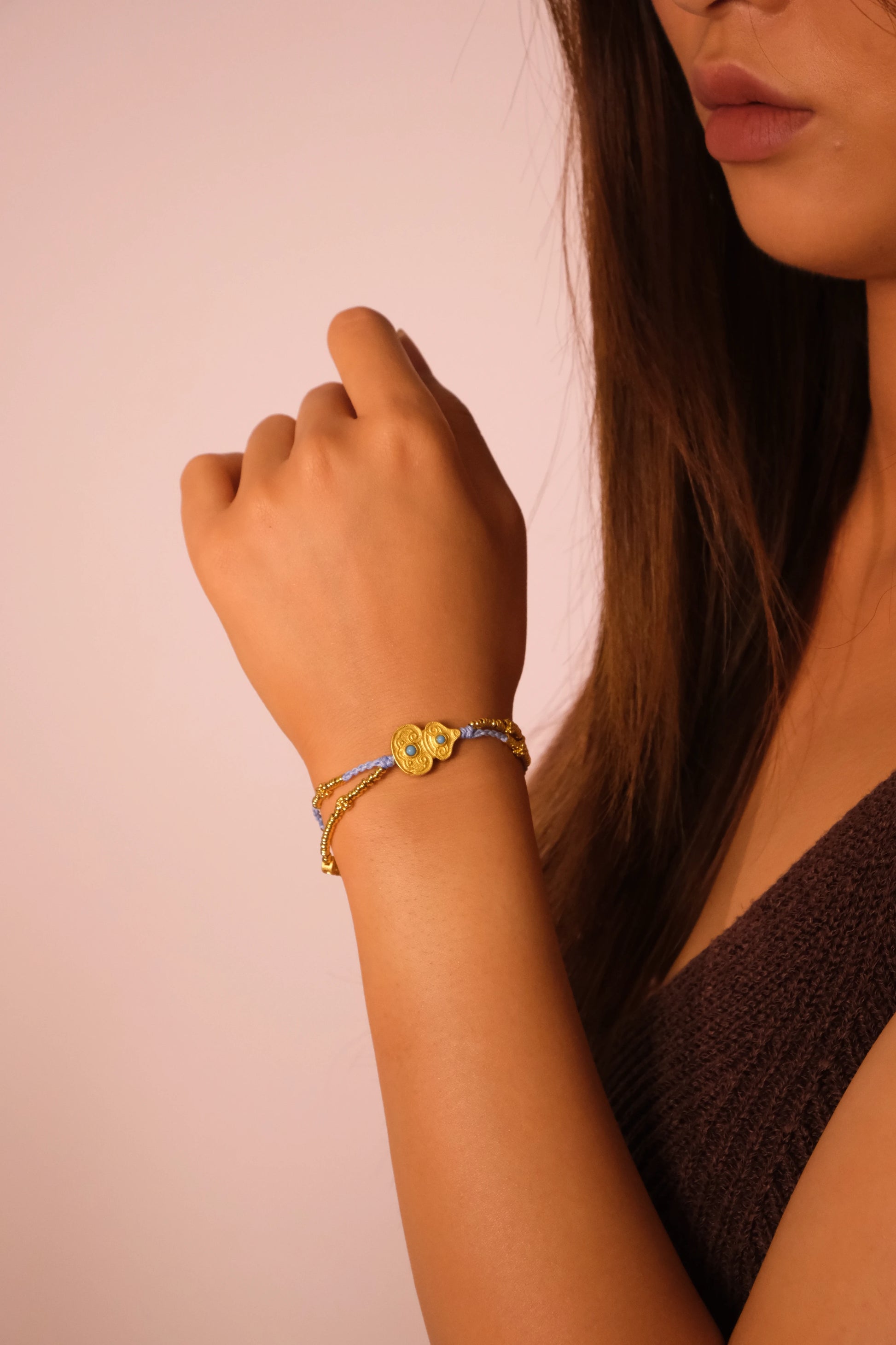 Close-up of a woman's arm wearing a bicolor bracelet on a pink background. Handmade bracelet with gold beads and blue woven cord, featuring a double-circle gold charm with two turquoise.