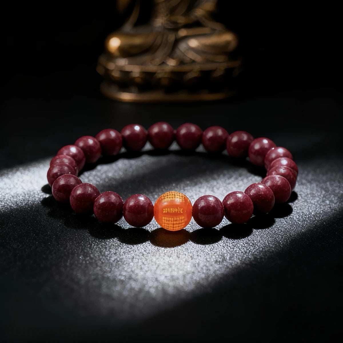 A red bead bracelet with one orange text-engraved bead, resting on a dark surface with a Buddha statue in the background.