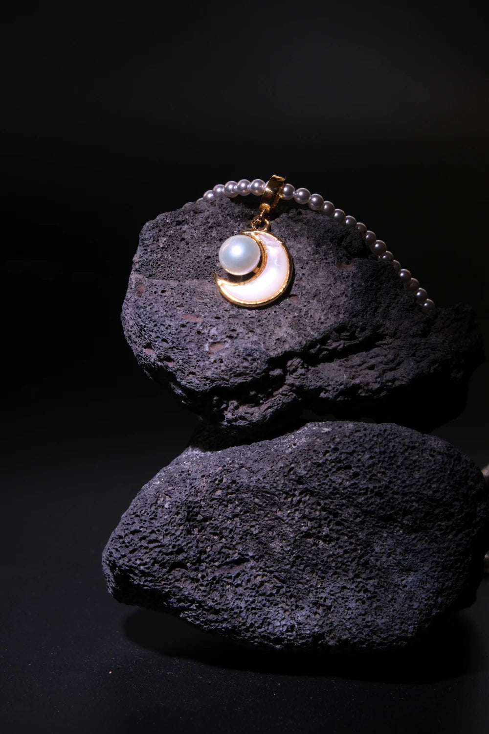 Handcrafted Tibetan Moonlight Pendant Necklace featuring a luminous mother-of-pearl pendant and pearls, showcased on volcanic rock with a dark background and focused lighting, associated with inner peace and feminine energy.